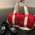 Load image into Gallery viewer, Red duffel bag with white handles and a black and blue design, next to black shoes on a dark surface.
