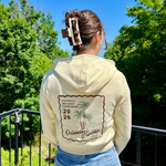 Load image into Gallery viewer, Person wearing a cream hoodie with a palm tree design, standing on a balcony with trees and blue sky in the background.
