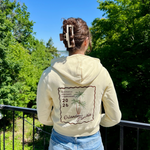 Load image into Gallery viewer, Person wearing a cream hoodie with a palm tree design, standing outdoors with trees and blue sky in the background.
