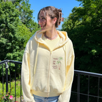 Charger l'image dans la galerie, Person wearing a light yellow hoodie with a graphic design outdoors on a sunny day.
