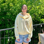 Charger l'image dans la galerie, Person wearing a light-colored hoodie with text and graphics, standing outdoors with green trees in the background.
