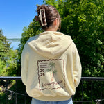 Charger l'image dans la galerie, Person wearing a cream hoodie with a palm tree design and text, standing outdoors with trees and water in the background.
