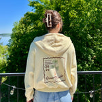Charger l'image dans la galerie, Person wearing a cream-colored hoodie with a palm tree design and text, standing outdoors with trees and a lake in the background.
