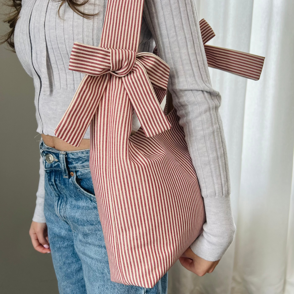 Person holding a pink and white striped bag with a large bow, wearing a light gray sweater and blue jeans.