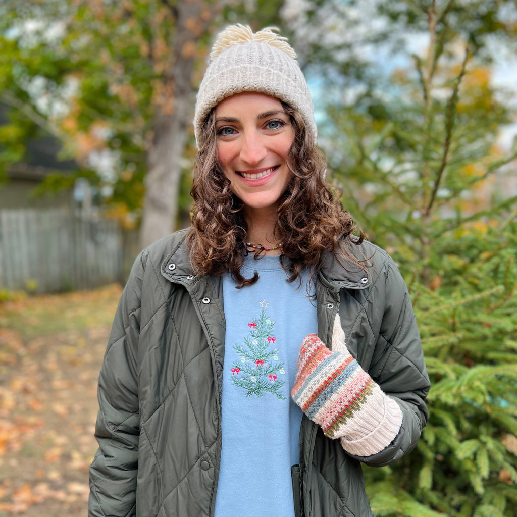 Woman wearing a green jacket, light blue shirt with a tree design, white beanie, and colorful mittens outdoors.