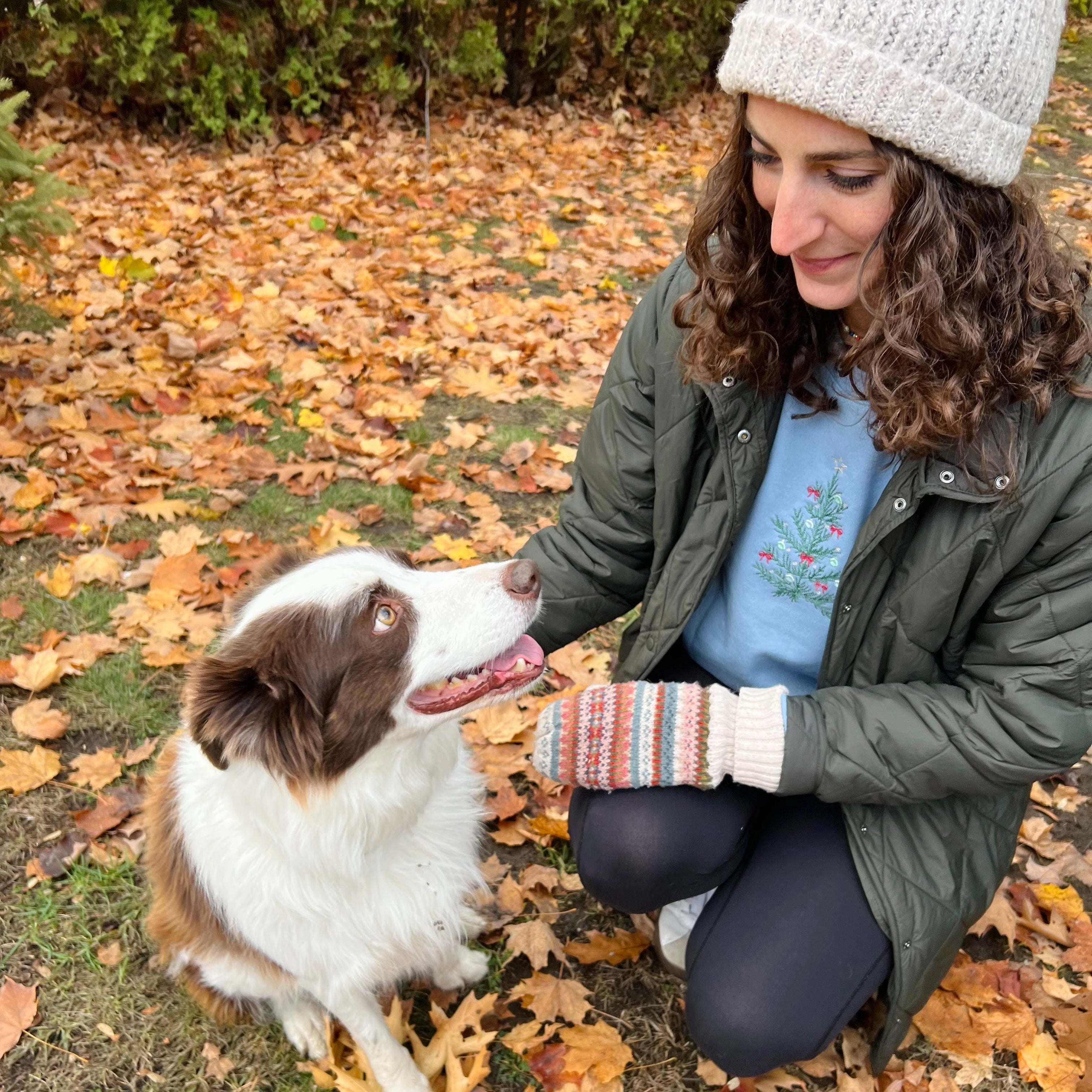 Woman in a green jacket and white beanie interacting with a brown and white dog on a leaf-covered ground.