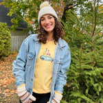 Load image into Gallery viewer, Person wearing a yellow shirt with a graphic design, denim jacket, and white beanie standing in front of a tree.
