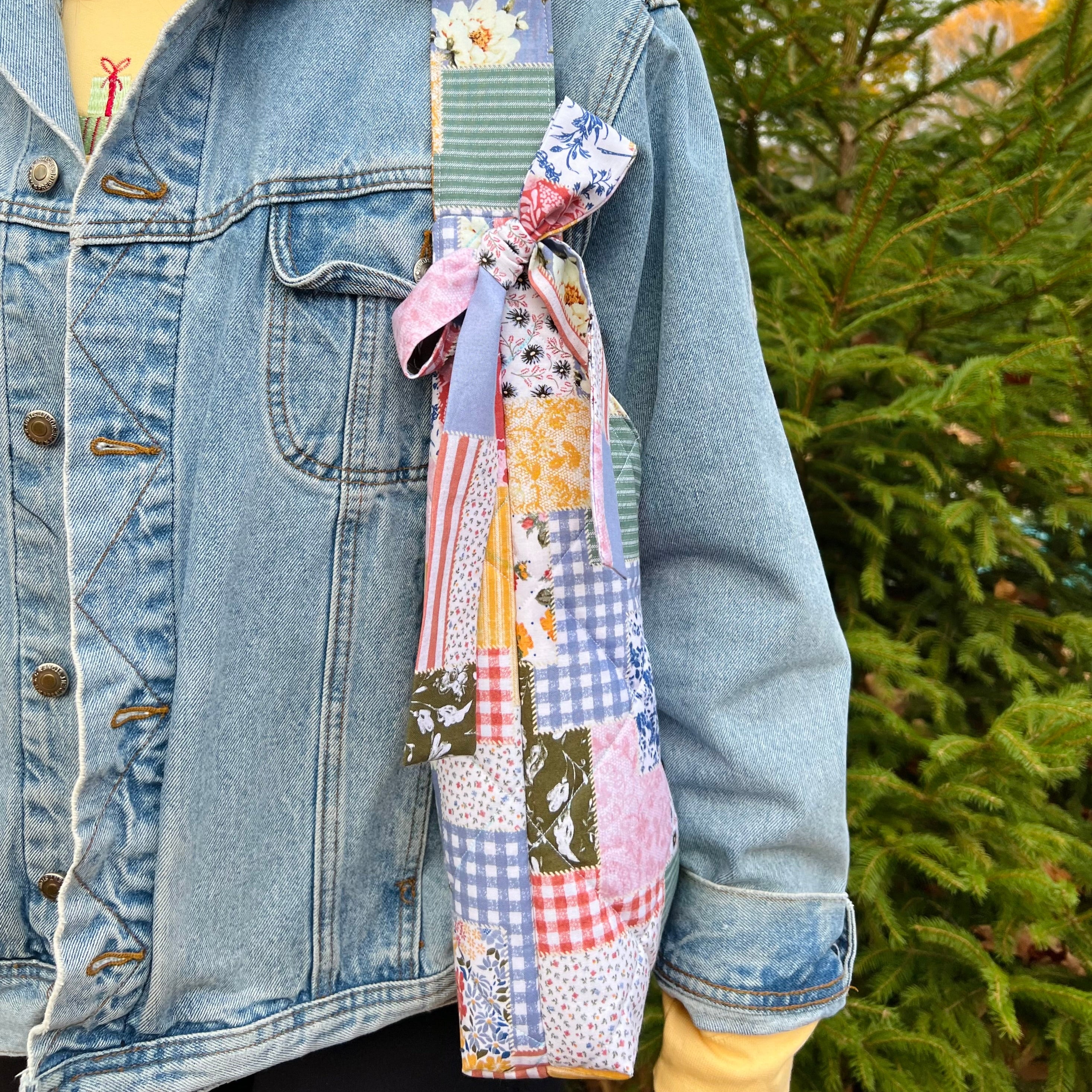 Person wearing a denim jacket with a patchwork quilt draped over their shoulder, standing outdoors.