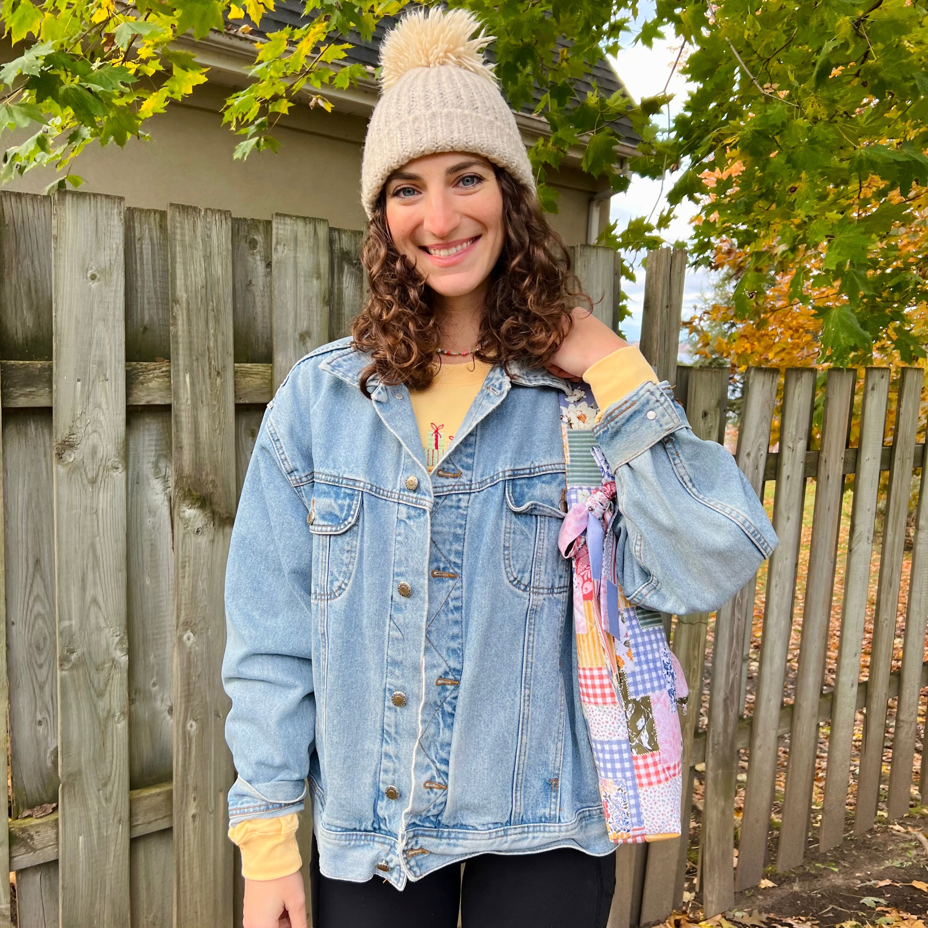 Woman wearing a denim jacket and beanie outdoors with a wooden fence and trees in the background